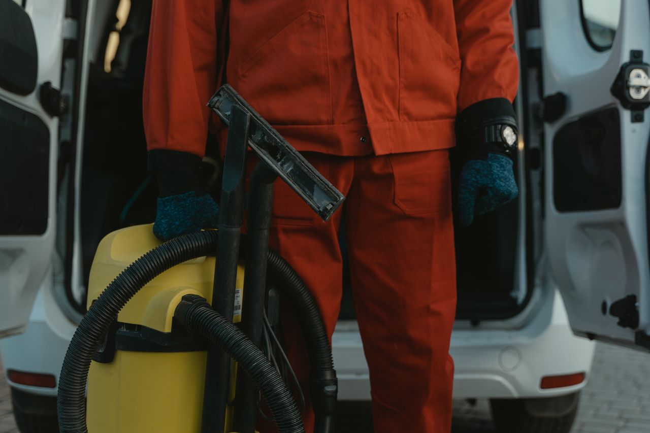 Un agent d’entretien professionnel en uniforme rouge, tenant un aspirateur près d’un véhicule utilitaire, prêt à intervenir.