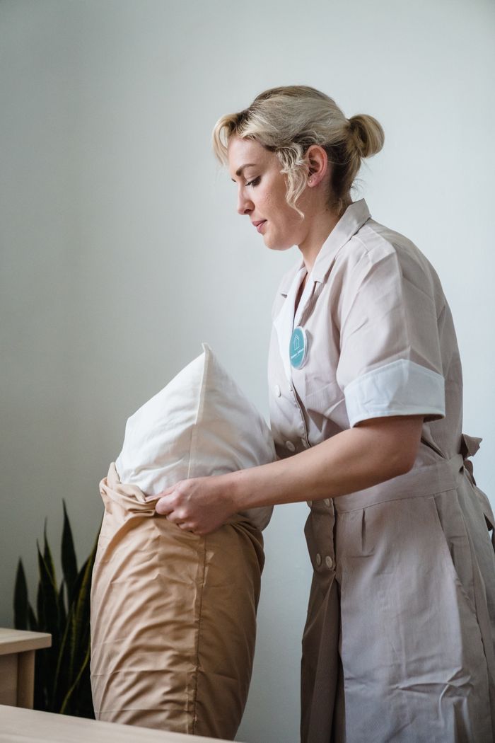 our-story Hotel maid in uniform arranging pillows in a neatly styled guest room.