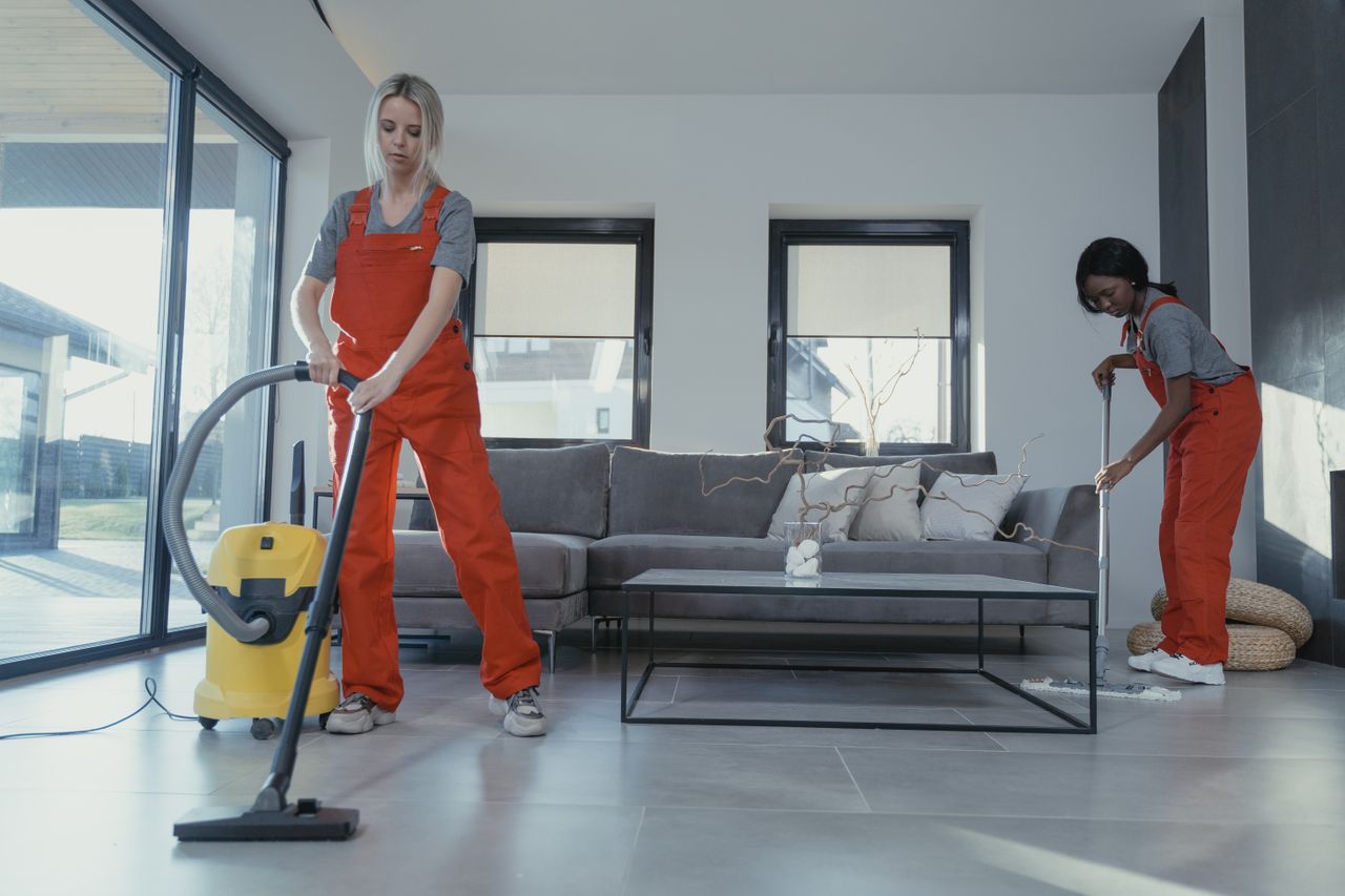 about-img-01 Two women in red overalls vacuuming and sweeping a modern living room.